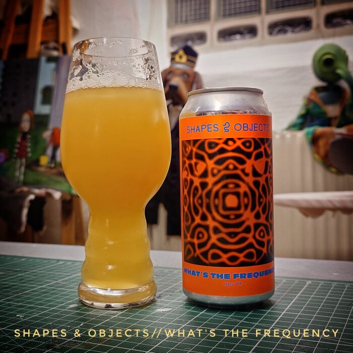 Ölglas med gyllene öl bredvid en burk med etikettexten "SHAPES & OBJECTS/WHAT'S THE FREQUENCY". Bakgrund med konstverk och diverse föremål.