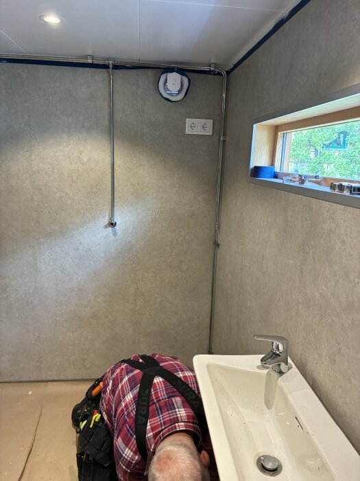 Person som installerar tvättställsblandare i nyrenoverat badrum med grå väggar och liten rektangulär fönsteröppning.