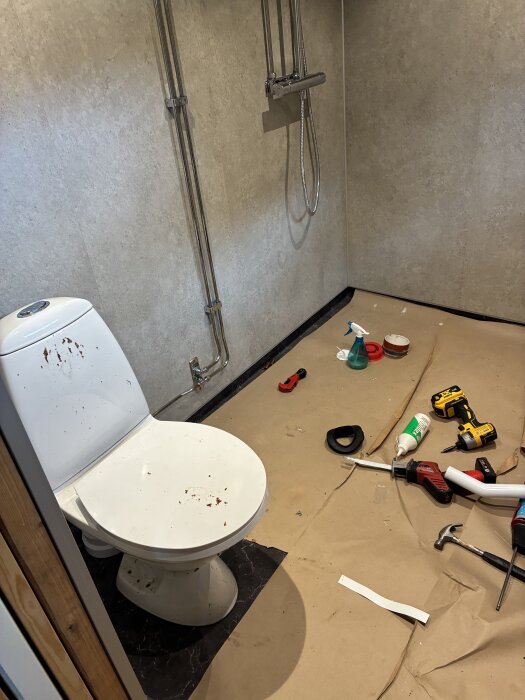 Toalettstol med smuts på sitsen, omgiven av verktyg och material på golvet, bredvid en duschblandare på en vägg med våtrumsskivor.