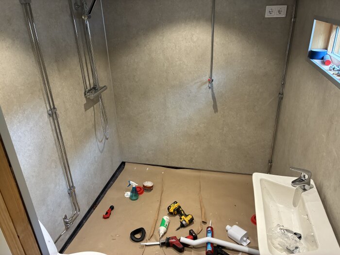 Badrum under renovering med grå våtrumsskivor, en Gustavsberg duschblandare, verktyg och material på golvet, handfat med en Hansgrohe kran.