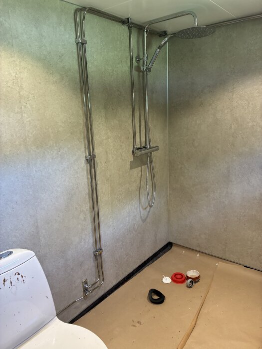 Badrum med duschblandare, grå våtrumsskivor och toalettstol; byggmaterial på golvet.