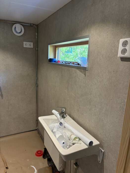 Tvättställ med Hansgrohe blandare, små fönster ovanför med utsikt, ventil och eluttag på väggen i oinrett badrum.