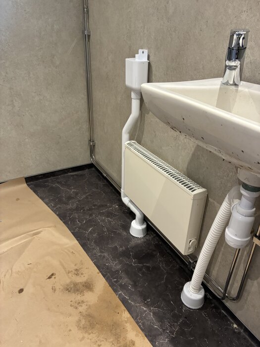 Tvättställ och radiator i ett nyrenoverat badrum med marmorerad plastmatta och rörinstallationer. Pappersskydd på golvet syns.