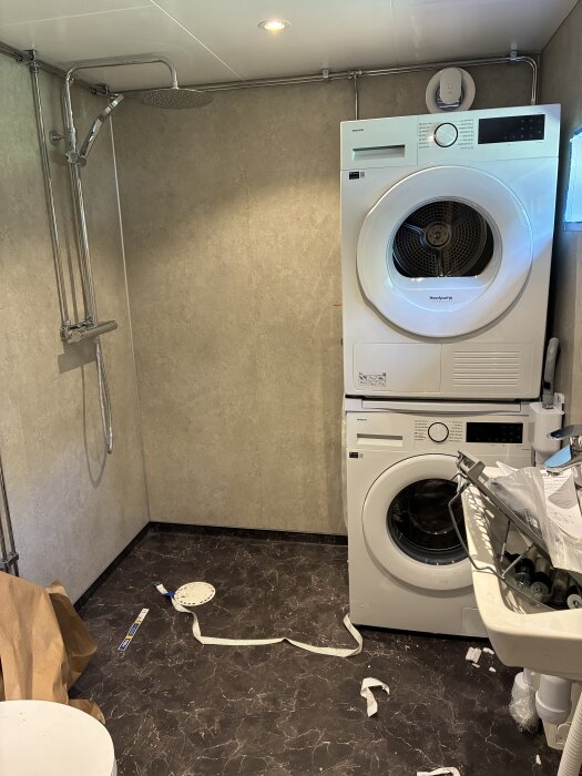 Badrum med Samsung tvättmaskin och torktumlare, duschutrustning installerad, Fibo våtrumsskivor och plastmatta, ännu osopade rester på golvet.