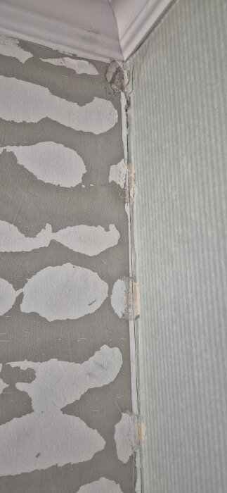 Närbild på ett hörn av ett rum med avslitet grått tapet och skadad vägg. Vitt tak med en list syns upptill.