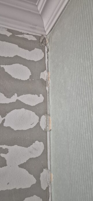 Hörn av rum med sprucken tapet och ojämn vägg, synlig list och skada i tapetskarv, delvis avskalad färg.