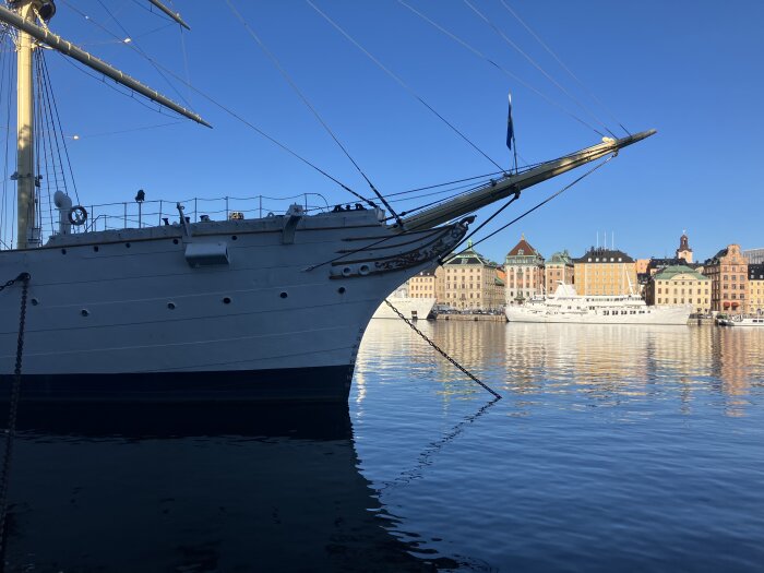 Segelbåts bog vid kaj, klart vatten, stadshus och färgglada byggnader i bakgrunden under en klarblå himmel.