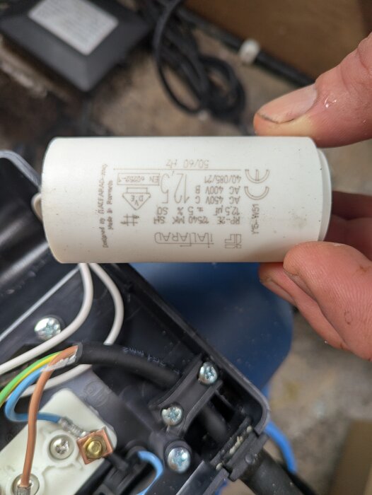 En hand håller en vit kondensator märkt med "12.5 450V" nära en elektrisk koppling med sladdar och skruvar.