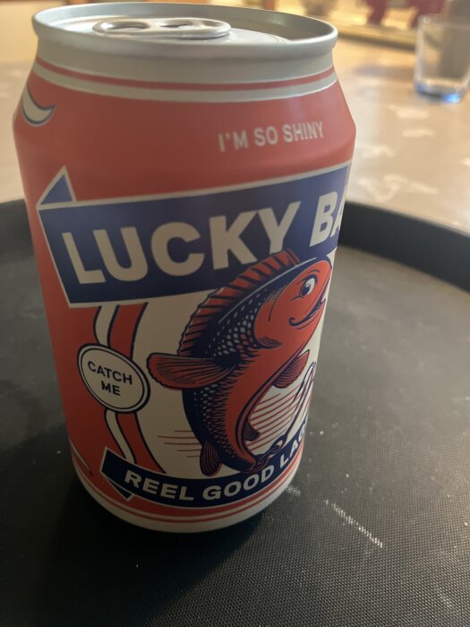 En ölburk med texten "Lucky Bay", en orange fisk och humoristiska fraser på etiketten, står på ett bord.