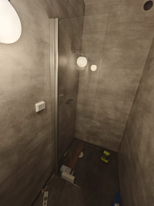 Badrum med mörk matta, glasvägg och lampa. Verktyg och material på golvet visar ett pågående renoveringsprojekt.