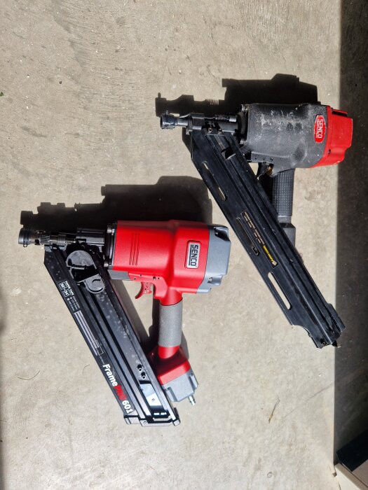 Två spikpistoler liggande på marken; en röd ny och en gammal, båda av märket Senco.