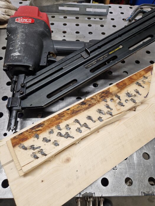 Nagelpistol på ett metallbord bredvid träbitar med fastskjutna korta spikar, använd i ett renoveringsprojekt.
