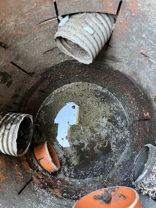 Dagvattenbrunn med korta och långa rör, delvis fylld med vatten och smuts. Ett böjt rör sticker ut och betongbotten syns delvis.