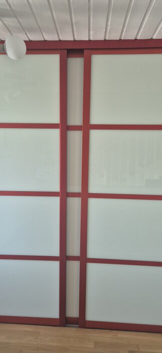 Röda metallramar runt frostade glasdörrar inomhus, synlig takvinkel och trägolv.