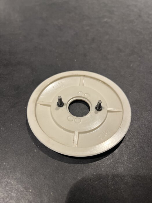 Rund täckring i bakelit med två fästskruvar, avsedda för strömställare, liggande på en mörk yta.