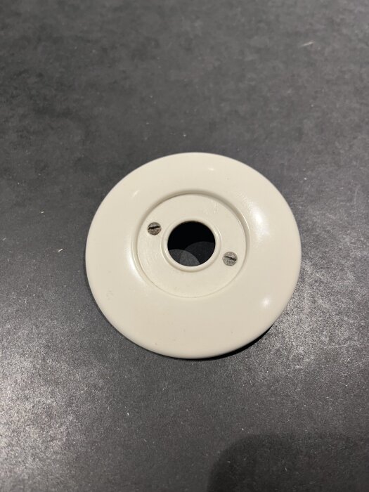 Vit bakelitströmbrytarring med två synliga fästskruvar på mörk yta, i behov av längre skruvar.