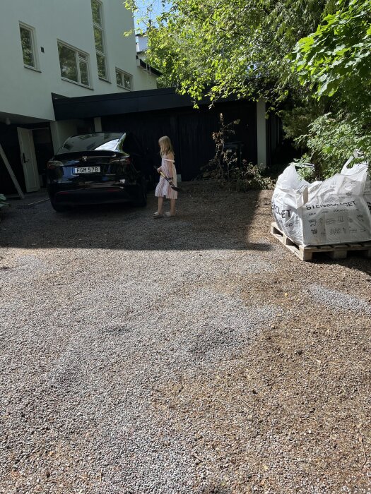 Flicka står på grusuppfart bredvid en bil, omgiven av löv och barr. Byggsäckar vid sidan och hus i bakgrunden.