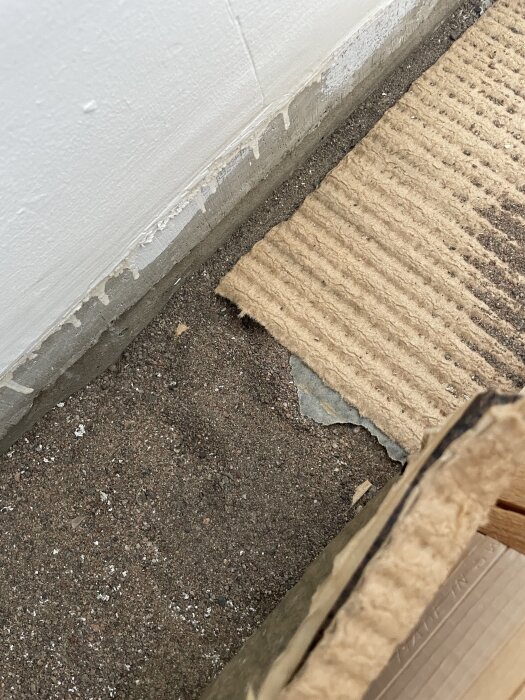 Gammal parkett rivs upp, under finns papp och lager av sand. Väggens nederdel syns, yta förbereds för nytt parkettgolv i vardagsrum.