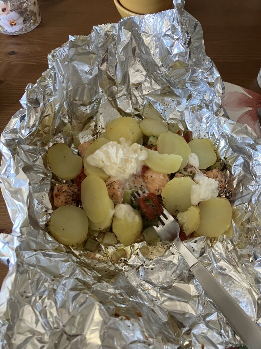 Foliepaket pÄ grillen med laxkuber, skivad potatis, purjolök, tomat och VÀsterbottenoströra. En gaffel syns pÄ sidan.