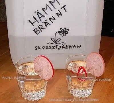 Två glas med dryck garnityrerade med korvskivor, framför en behållare märkt "Hämm Brännt Skogsstjärnan".