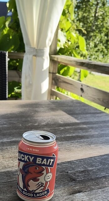 En ölburk med etiketten "Lucky Bait" står på ett träbord med grönska i bakgrunden.