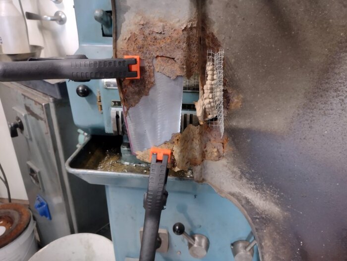 Reparation av en rostskadad framskärm på en bil, med metallplåt fastklämd på plats för svetsning. Rost och nätförstärkning syns tydligt.