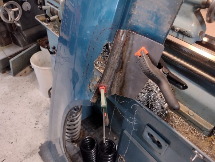 Blå framskärm på Volvo med ny plåtbit fastklämd, omgiven av metallspån och verktyg i verkstadsmiljö.