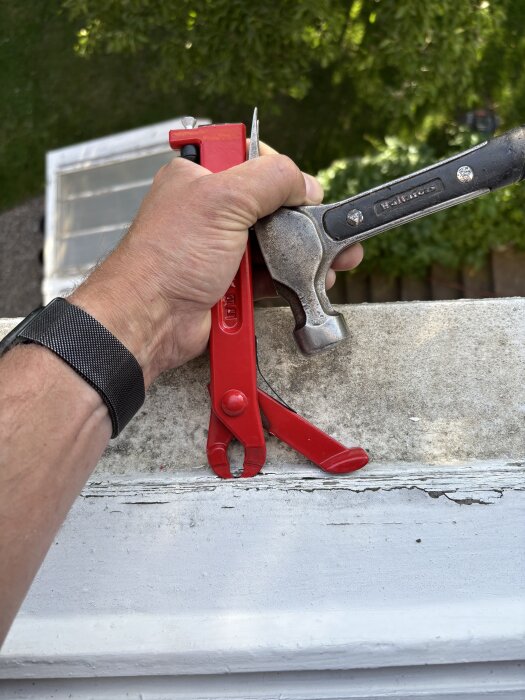 Hand håller en Hultafors hammare och en röd tång på en betongkant utomhus.