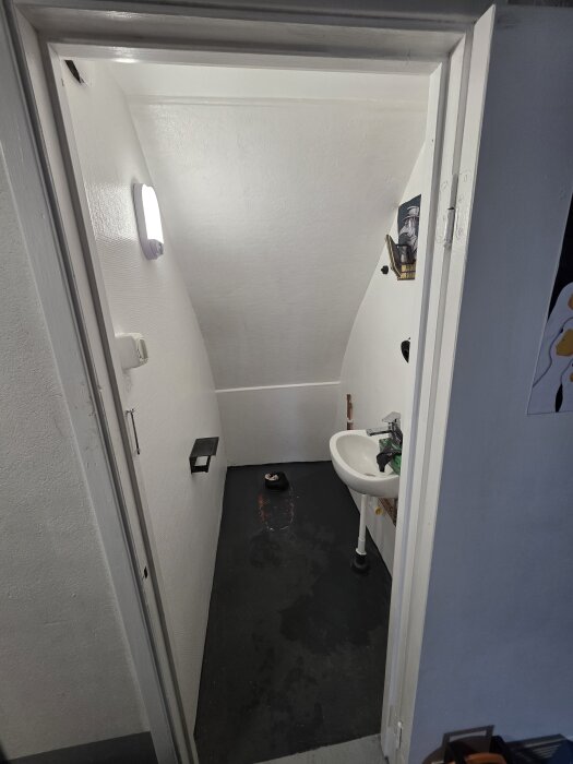 Toalett i källare under renovering med ett frilagt hål i golvet, svart golv, vit vägg, handfat och en hylla med prydnad ovanför.