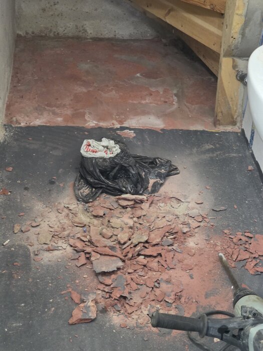 Bilad upp golv i källartoalett med krossade betongbitar på golvet och ett verktyg bredvid. Svart plastpåse ligger ovanpå betongen.