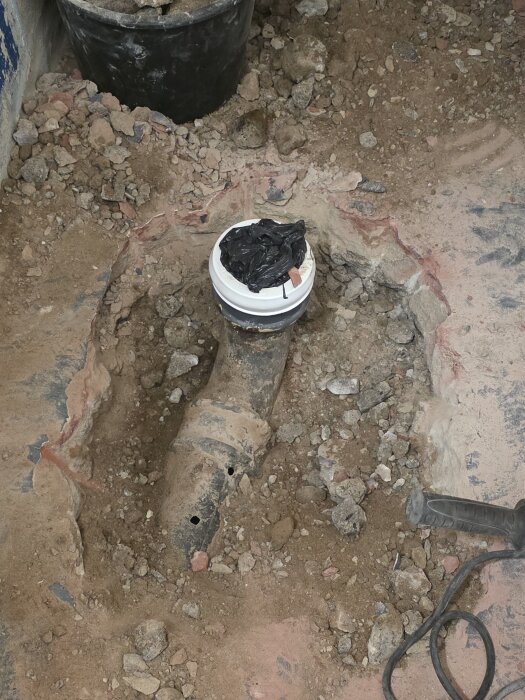 Bilat golv med avloppsrör i källare under renovering. Plasttäckning och stenrester syns, verktyg till höger.