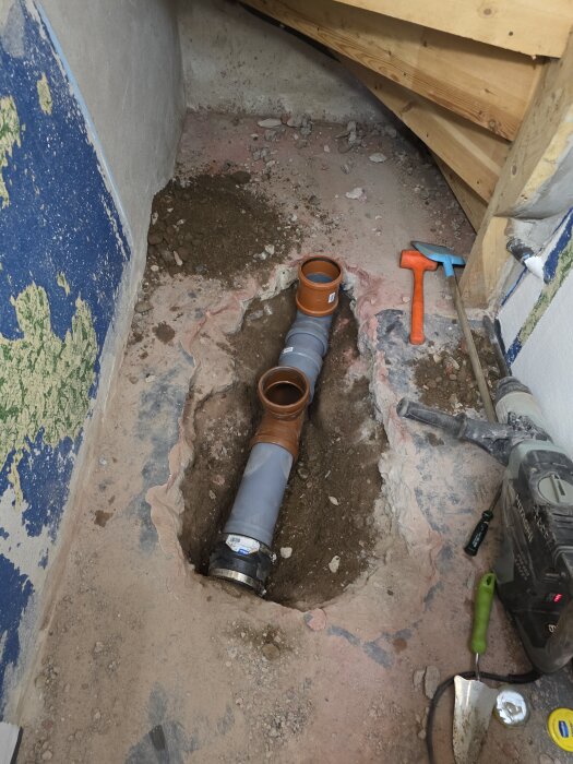 Uppgrävd golvyta i källare med synliga avloppsrör och verktyg som hammare och spade, del av renoveringsprojekt för badrum.