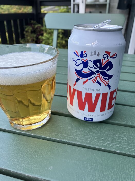 Ölglas och en öppen ölburk på ett grönmålat trädgårdsbord.
