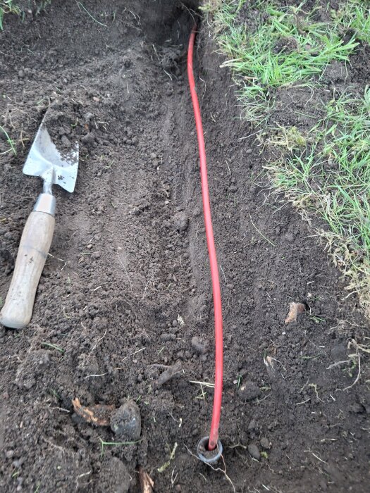 Röd kabel i grävd jord, delvis täckt av röd skyddsslang, bredvid en liten spade och gräsmatta; fiberkabel på 3 cm djup.