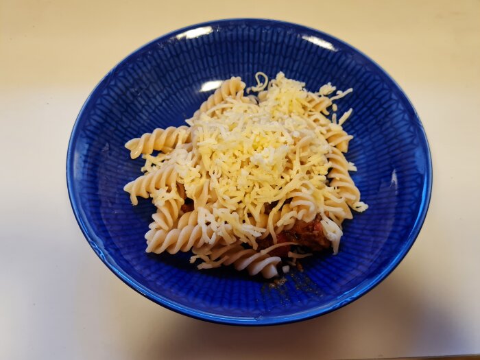 En blå skål med bönpasta toppad med riven tacoost, med pastasås dold under fusillin.