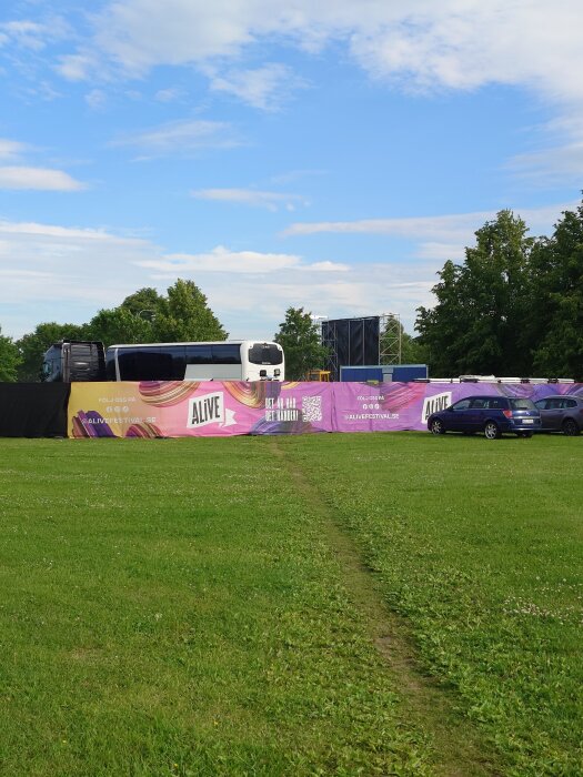 Gräsfält med en färgglad festivalstaket, en buss och bilar i bakgrunden under en blå himmel.