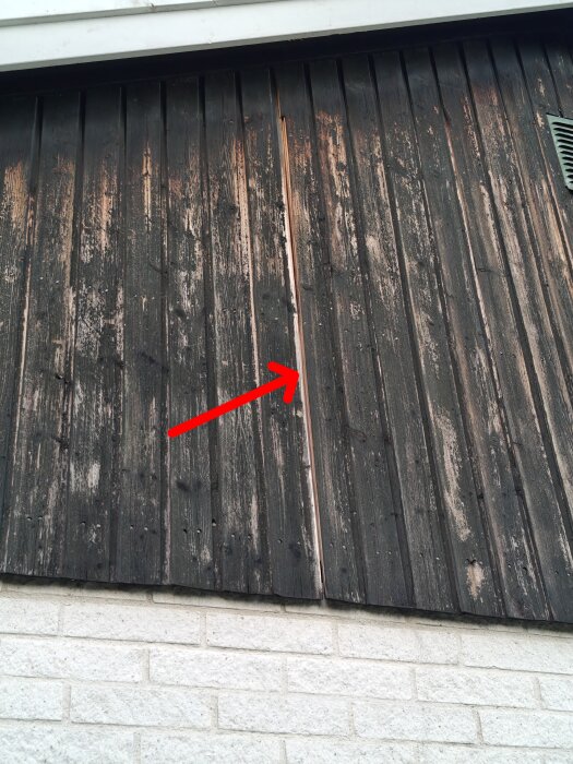 Träpanel på husvägg med sliten färg och en tydlig spricka där underliggande plywood är synlig, markerat med röd pil.
