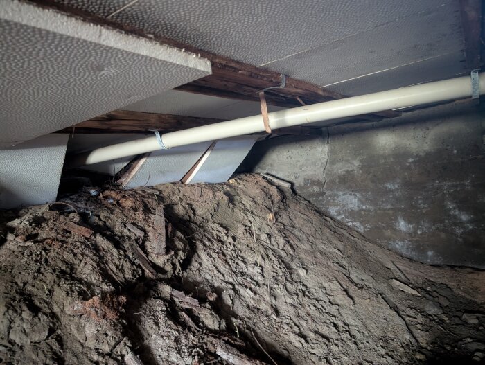 Innerbild av ett gammalt hus med synliga rör och jordhög, visar isolering och möjlig fuktskada i källarutrymme.