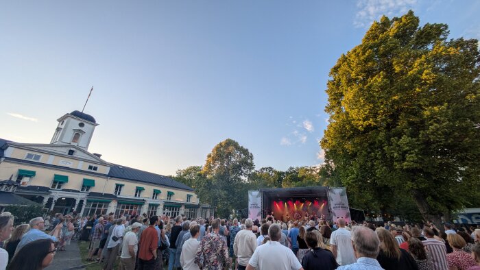 Publik framför en scen vid en utomhuskonsert i Brunnsparken, Söderköping, med höga träd och en byggnad i bakgrunden under en klar himmel.