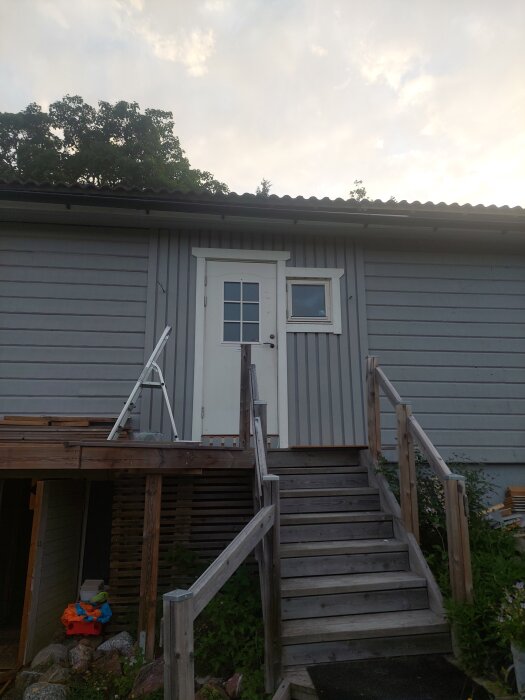 Gråmålad huspanel och foder, trappa leder upp till dörr. Vitt fönster bredvid dörren, himmel och träd i bakgrunden. Brusigt vid entrén.