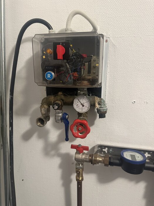 Pumpsystem med rör och ventiler i en brunnsanläggning, inklusive tryckmätare och kontrollbox på väggen.