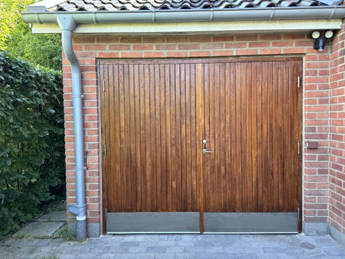 Brun trägarageport med två dörrar, rött tegelhus och ett grått stuprör på vänster sida, omgiven av gröna buskar.