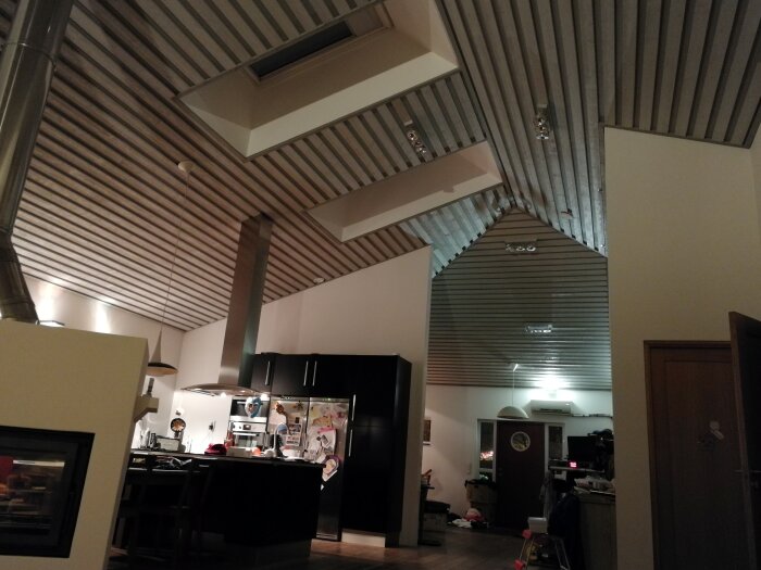 Modernt kök med högt tak, takfönster och synliga takbjälkar. Köket har en köksö, svart skåpinredning och en spis under en rostfri fläkt.