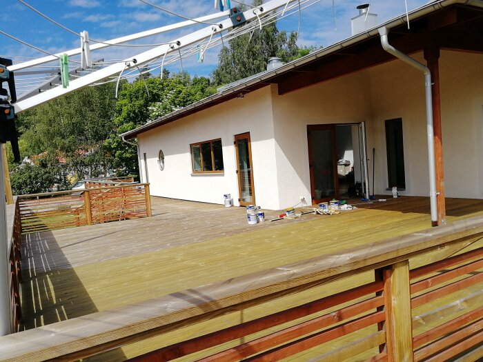 Trädäck med tillhörande vitputsat hus och öppna dörrar, omgiven av grönska. Målarverktyg syns på trädäcket.