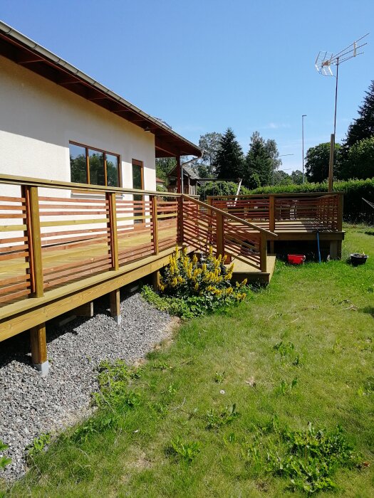 Träaltan med räcke vid sidan av ett hus med vita väggar och stora fönster, gräsmatta och antenn i bakgrunden.