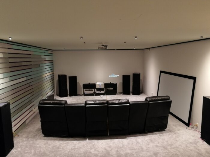 Hemmakino med svarta fåtöljer, stor filmduk på golvet, högtalare och projektor i ett rum med randig vägg.