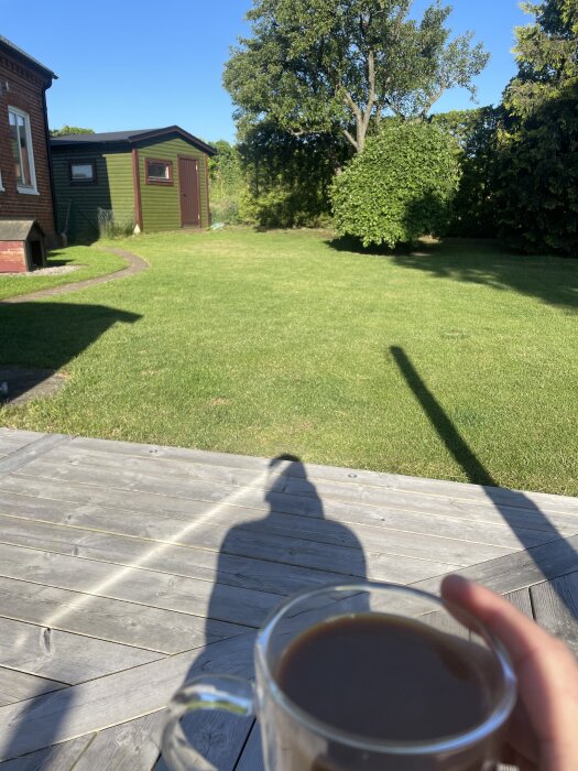 Hand håller en kaffemugg på en solig träaltan, sedd med grönt gräs och träd i bakgrunden. En grön byggnad syns också.