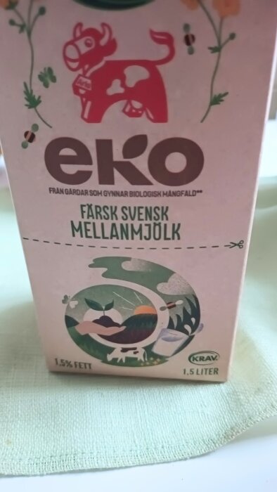 Ekologiskt mjölkpaket med illustrationer och texten "Färsk Svensk Mellanmjölk". Mjölkpaketet har orsakat spill på bordet.