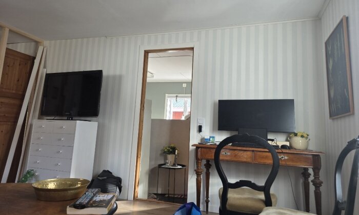 Rum med vit- och grårandig tapet, entrédörr utan dörrkarm, TV på byrå och skrivbord med dator samt stol.