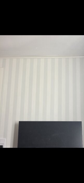 Randig tapet i ljusa nyanser på en vägg med en del av ett mörkt möbelstycke nedanför.